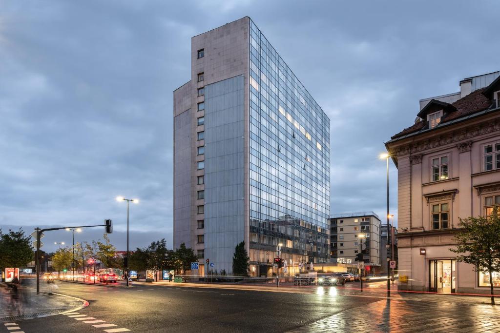 Modern high-rise Exe Lev hotel in Ljubljana at dusk, with a glass-and-stone facade reflecting the cloudy sky, illuminated streetlights, and light trails from passing cars on the wet street in front.