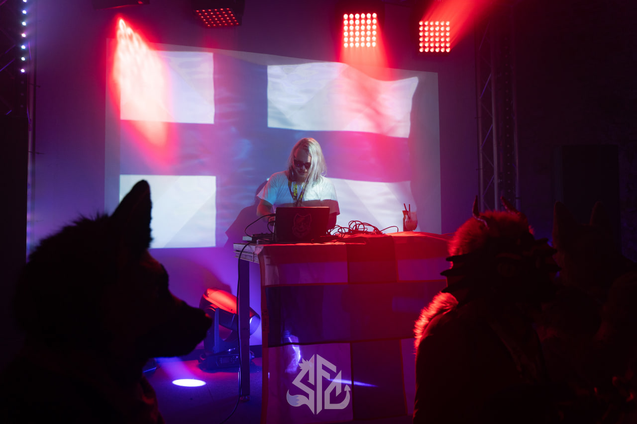 DJ performing at SloFluffCon behind a booth bearing the SFC logo, with the Finnish flag projected on the back wall and two fursuiters silhouetted in the foreground watching the set.