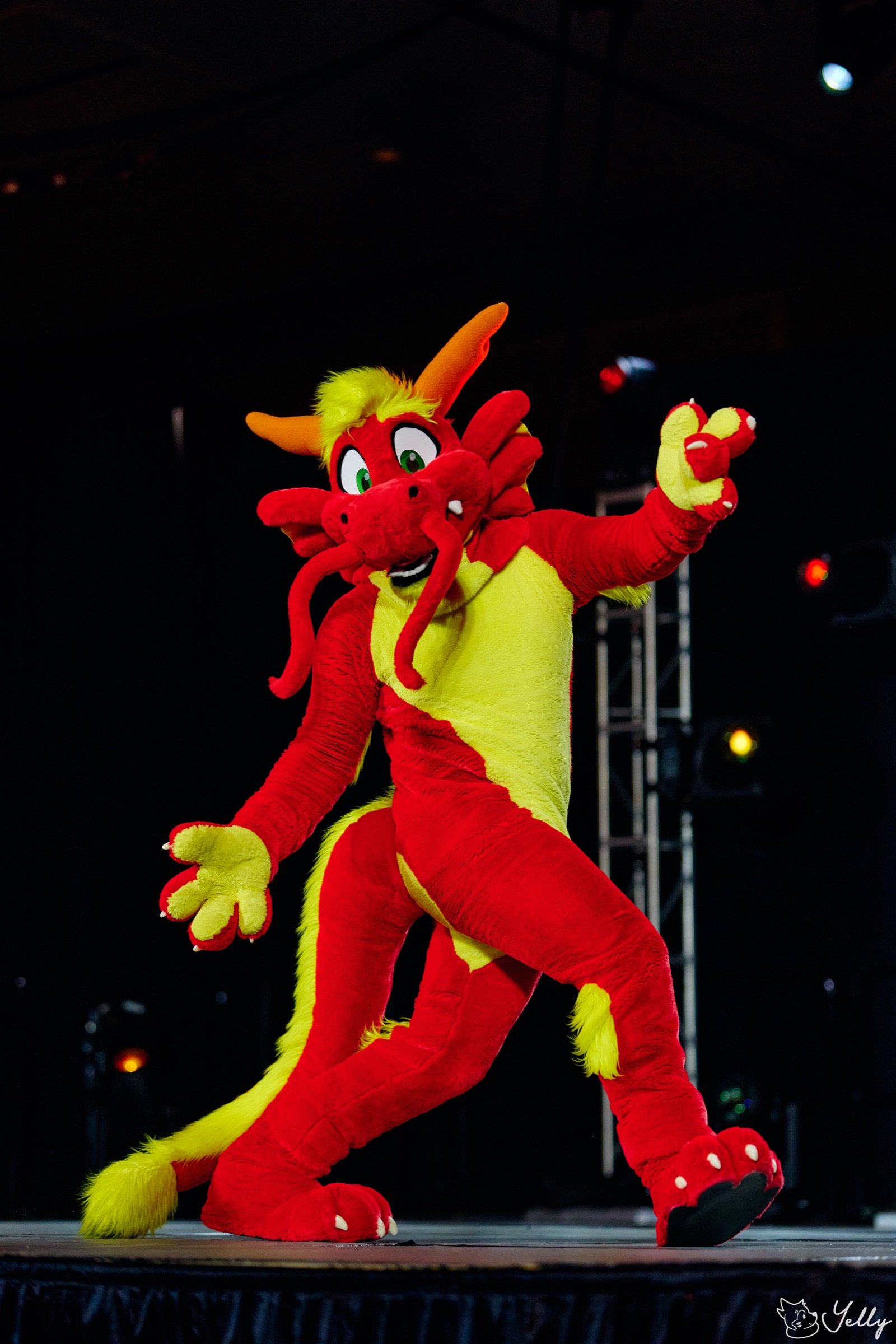 Ronnie Noodles, a red and yellow dragon fursuiter, mid-dance on a stage under truss lighting