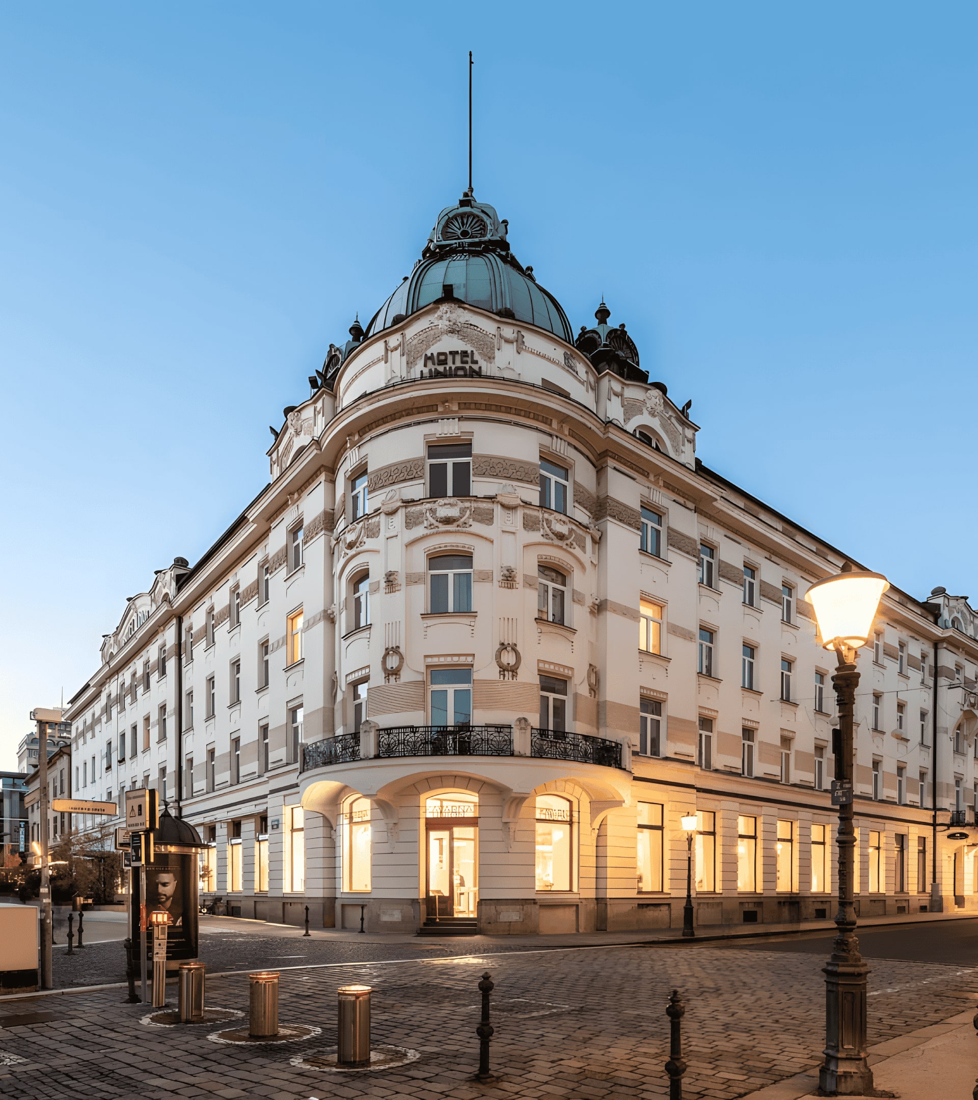 Corner view of the Hotel Union building in Ljubljana, Slovenia, illuminated at dusk, with ornate architectural details, a copper-green dome on top with the word "HOTEL" visible, a cobblestone street, streetlights, and flags hanging across the street.