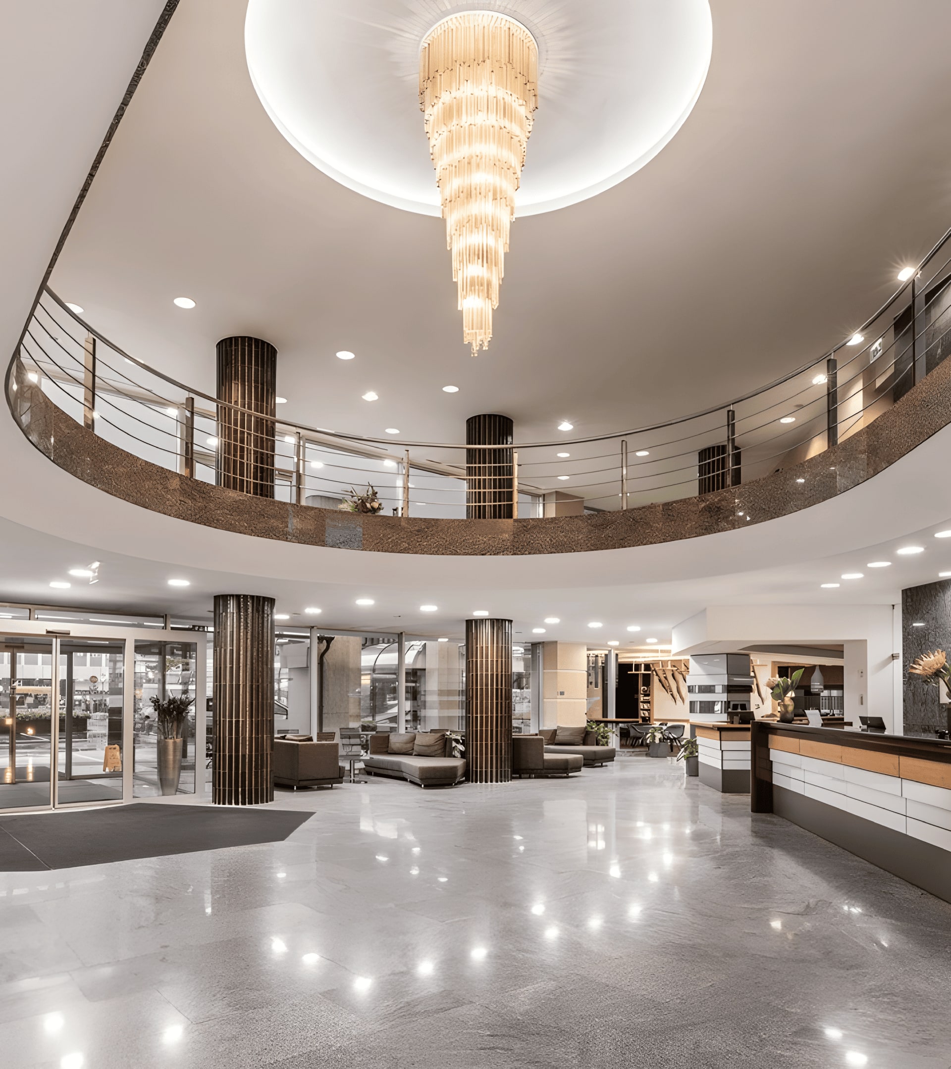A brightly lit hotel lobby with a grand, tiered chandelier hanging from a recessed circular ceiling. The lobby has a polished granite floor reflecting the light, and a curved mezzanine level with a metal railing. The front desk, made of wood, sits under a large green marble wall. Multiple dark cylindrical columns with golden brown tile wraps line the lobby floor. Large glass doors lead to the outside, visible through the windows.